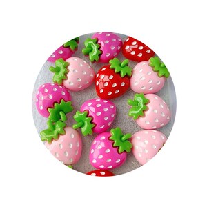 May include: A collection of strawberry-shaped embellishments in various shades of pink and red, with green leafy tops. The strawberries have a glossy finish and are arranged in a circular pattern. The image is set against a light gray background.