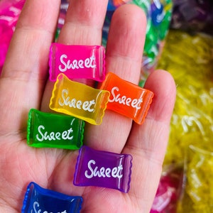 May include: Six colorful plastic candy wrappers with the word "Sweet" printed in white on each. The wrappers are pink, yellow, orange, green, purple, and blue.