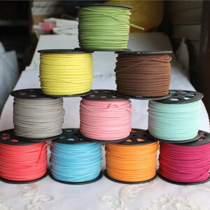 Op de afbeelding: Tien spoelen met gekleurde suède koord, elk gewikkeld rond een zwarte plastic spoel. De kleuren omvatten geel, groen, bruin, grijs, roze, lichtblauw, oranje en rood.
