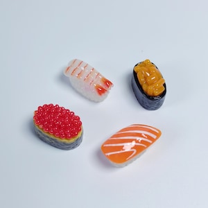 May include: Four miniature sushi pieces including a shrimp nigiri, a salmon nigiri, a gunkan maki topped with bright red fish roe, and a gunkan maki topped with yellow pieces, all featuring a glossy, realistic finish and textured details.