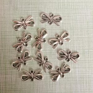 Pode incluir: Doze pingentes de metal em forma de laço, de cor prateada, com um laço na parte superior para prender em joias ou outros trabalhos manuais.
