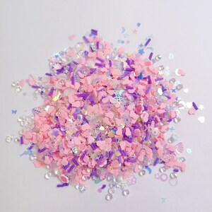Puede incluir: Un montón de suministros de manualidades en colores pastel, que incluyen formas de flores y conejos rosas, fideos morados y lentejuelas iridiscentes. La mezcla también presenta estrellas, lunas y otras formas pequeñas. Los artículos están esparcidos sobre una superficie blanca.