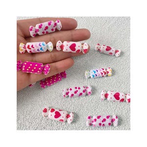 May include: A collection of miniature candy-shaped embellishments. The candies are white with pink, red, and blue heart and triangle patterns. Some candies are pink with gold foil accents. These are likely used for crafts or decorations.