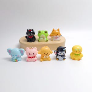 Könnte beinhalten: Eine Sammlung von neun Miniatur-Tierfiguren, darunter eine schwarze Katze, ein grüner Frosch, ein orangefarbener Fuchs, ein blauer Elefant, eine rosa Katze, ein gelber Hund, ein Pinguin und eine gelbe Ente. Die kleinen, glatten Figuren sind in zwei Reihen angeordnet.