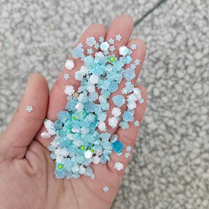 May include: A hand holding a variety of blue and white flower-shaped embellishments. The embellishments are made of plastic and have a glossy finish.