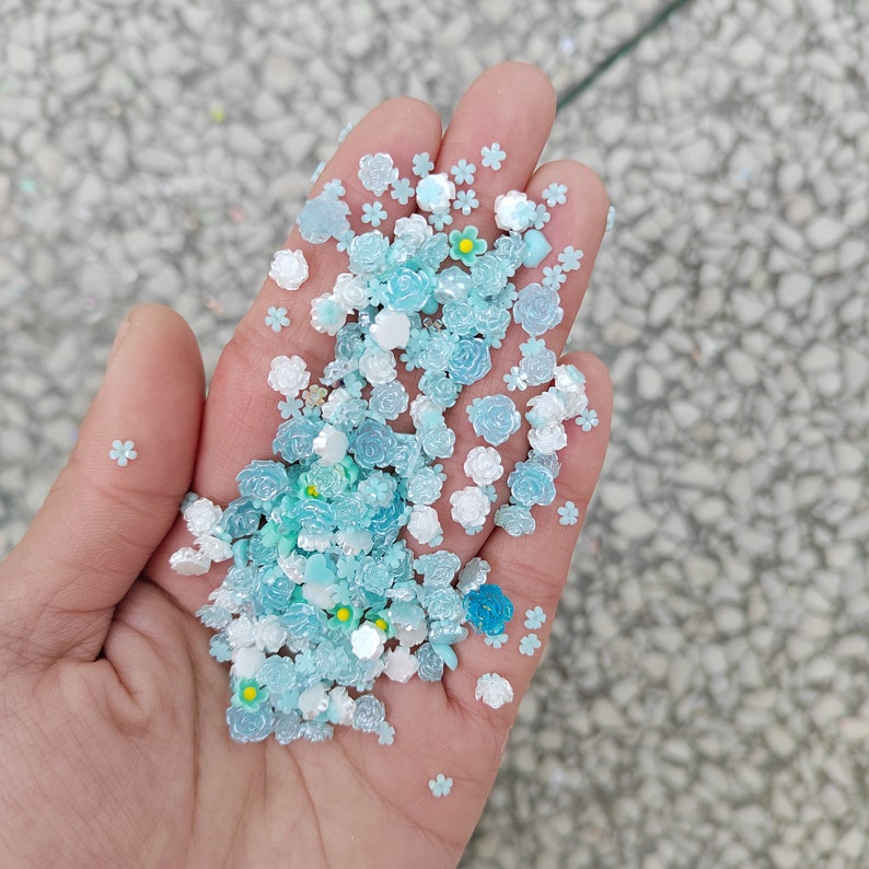 May include: A hand holding a variety of blue and white flower-shaped embellishments. The embellishments are made of plastic and have a glossy finish.