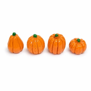 Puede incluir: Cuatro pequeñas figuras de calabaza de color naranja brillante dispuestas en fila. Cada calabaza presenta una forma distinta con crestas verticales y está rematada con un pequeño tallo verde liso. Las superficies son de color naranja vivo.