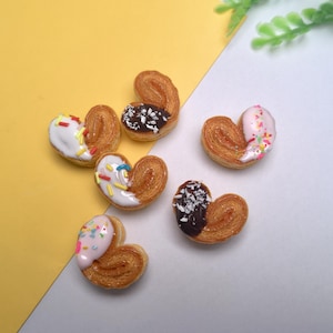 May include: An overhead shot of six heart-shaped pastries, each with a golden-brown, flaky exterior. Some are topped with white, pink, or chocolate icing, decorated with sprinkles. The pastries are arranged on a yellow and white surface.