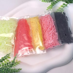 May include: Five clear plastic bags filled with colorful, shredded soap. The colors include light green, red, yellow, pink, and black. The bags are arranged in a row on a white surface, with green foliage in the background.