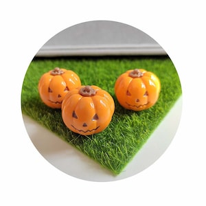 May include: Three miniature orange pumpkins with carved jack-o'-lantern faces, sitting on a bed of green artificial grass.