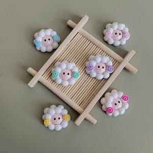 May include: Six white sheep-shaped resin embellishments with pink faces and colorful curly accents. The embellishments are arranged on a wooden tray with bamboo slats.