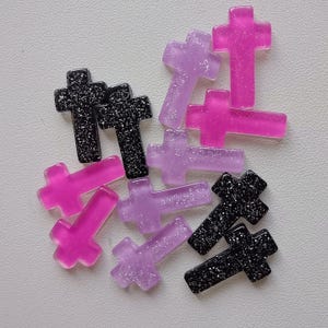 May include: A selection of decorative crosses in pink, purple, and black. Some crosses have glitter detailing. The crosses are made from a translucent material. The image shows a variety of cross designs.