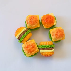 May include: A collection of miniature, colorful burger charms arranged in a circle. Each charm features a bun, patty, cheese, and lettuce, with vibrant orange, brown, yellow, and green hues. The charms are set against a plain white background.