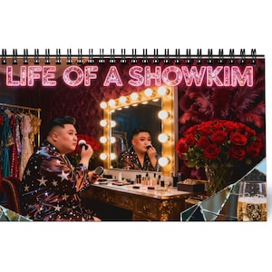 May include: A spiral-bound calendar featuring a person applying makeup in front of a lighted vanity mirror. The calendar's cover displays the text "THE LIFE OF A SHOWKIM" in pink neon. A bouquet of red roses and a glass of champagne are also visible.