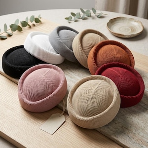 May include: A collection of nine round, brimless hats in various solid colors including black, white, gray, beige, orange, red, pink, and tan. Each hat features a dense, uniform knit texture and a rolled edge. A blank rectangular tag hangs from one hat.