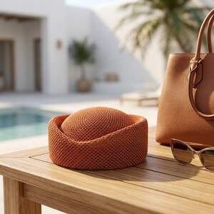 May include: A burnt orange woven hat with a rounded crown and a structured brim, accompanied by a tan leather handbag with dual handles and a pair of cat-eye sunglasses with gold-toned frames resting on a wooden surface.