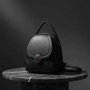 May include: A black leather backpack with a rounded flap and a small handle. The backpack features a circular clasp and white stitching details. The bag is sitting on a marble-topped table.