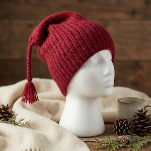 May include: A knitted red hat with a long tassel, displayed on a white mannequin head. The hat is made of a thick, textured yarn. The background includes a cream-colored scarf, pine cones, and a mug.