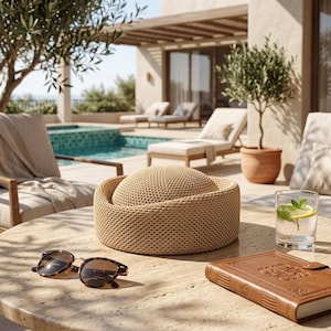 May include: A beige woven straw hat with a rounded crown and a wide, structured brim sits on a stone table next to a pair of brown tortoiseshell sunglasses and a brown leather-bound book, with a glass of water containing a lemon slice.