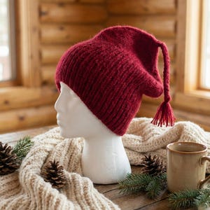 May include: A burgundy knit hat with a long, pointed top and tassel detail. The hat is displayed on a white mannequin head, with a cream-colored scarf, pinecones, and a mug on a wooden surface. The hat's design is a cozy, winter-themed accessory.