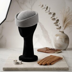 May include: A light gray pillbox hat with a textured, mesh-like surface pattern. The hat features a rounded crown and a structured, upright brim, displayed on a black mannequin head form alongside a pair of tan leather gloves and a silver cuff.