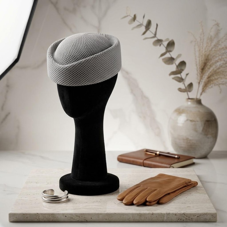 May include: A light gray pillbox hat with a textured, mesh-like surface pattern. The hat features a rounded crown and a structured, upright brim, displayed on a black mannequin head form alongside a pair of tan leather gloves and a silver cuff.