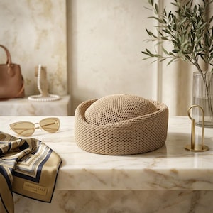 May include: A beige woven straw hat with a rounded crown and a structured brim. The hat features a tight, uniform knit texture throughout its surface, presenting a neutral tone and a circular silhouette that sits flat on a surface.