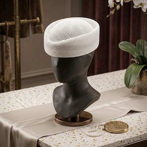 May include: A white pillbox hat with a structured, open-weave mesh texture. The hat features a rounded crown and a thick, solid band around the base, displayed on a dark, featureless mannequin head bust to showcase its form and design.