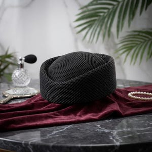 May include: A black pillbox hat featuring a structured, woven mesh texture. The hat has a rounded crown and a thick, uniform brim, showcasing a consistent grid-like pattern throughout its surface, presented as a singular, solid-colored accessory.