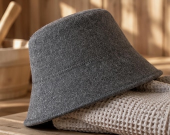 May include: A dark gray bucket hat with a textured, felt-like surface. The hat features a rounded crown and a downward-sloping brim, constructed with visible stitching along the base of the crown and the edge of the brim.