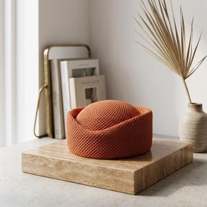 May include: A round, burnt orange knit hat with a structured, flat-topped crown and a wide, folded brim. The hat features a dense, uniform textured stitch pattern throughout, creating a consistent surface appearance across the entire object.