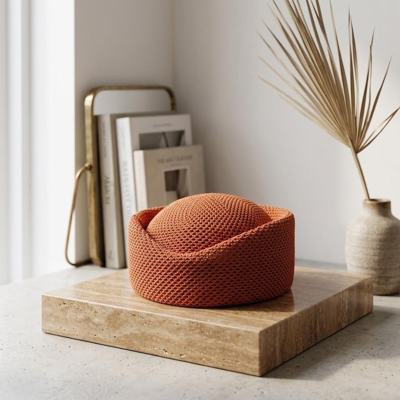 May include: A round, burnt orange knit hat with a structured, flat-topped crown and a wide, folded brim. The hat features a dense, uniform textured stitch pattern throughout, creating a consistent surface appearance across the entire object.