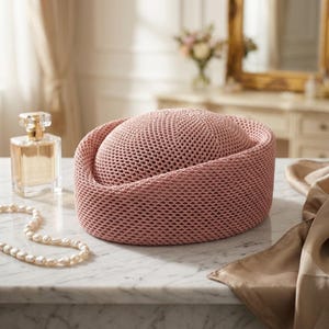 May include: A dusty rose pillbox hat featuring a structured, rounded crown and a wide, flat-topped brim. The hat is crafted from a fine, uniform mesh material with a consistent, textured weave throughout its entire surface, creating a solid appearance.