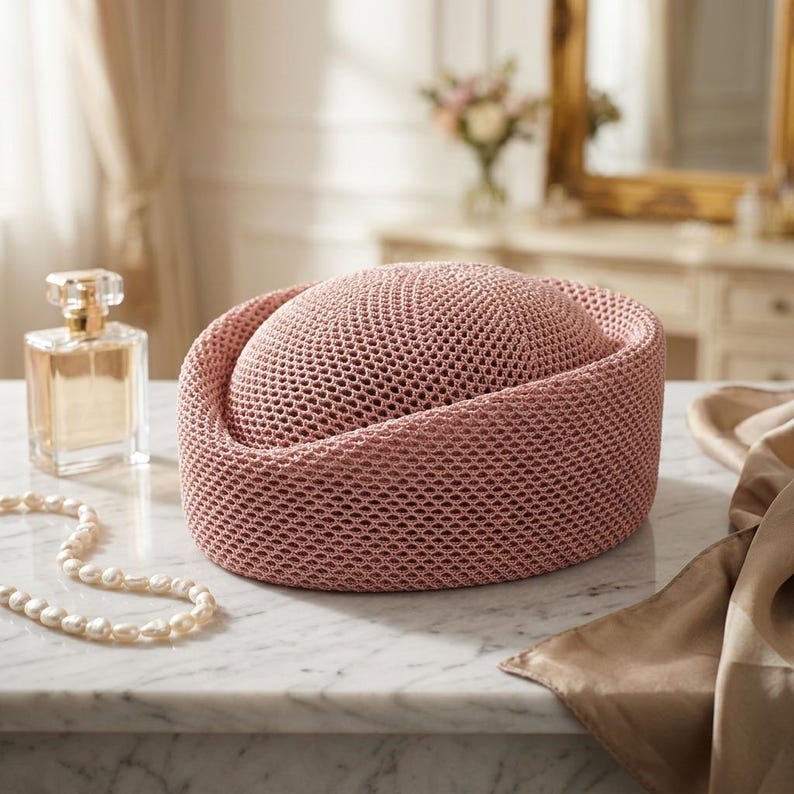 May include: A dusty rose pillbox hat featuring a structured, rounded crown and a wide, flat-topped brim. The hat is crafted from a fine, uniform mesh material with a consistent, textured weave throughout its entire surface, creating a solid appearance.