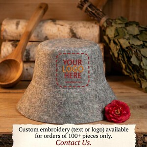 May include: A gray felt bucket hat with the text "YOUR LOGO HERE" embroidered on the front. A red rose is next to the hat. The hat is on a wooden surface. Custom embroidery is available for orders of 100+ pieces.