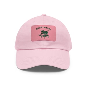 Puede incluir: Gorra de béisbol rosa claro con visera curva. La gorra presenta un parche rectangular con el texto "Chinga La Migra" y un gráfico verde de un gato sobre una mesa. El gato está junto a un rodillo con la palabra "ICE".