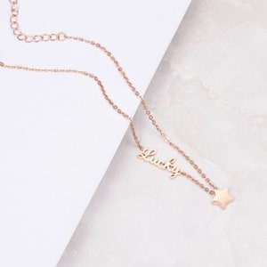 Puede incluir: Un collar de oro rosa con una cadena delicada y un colgante que dice "Lucky" con un peque&ntilde;o dije de estrella.