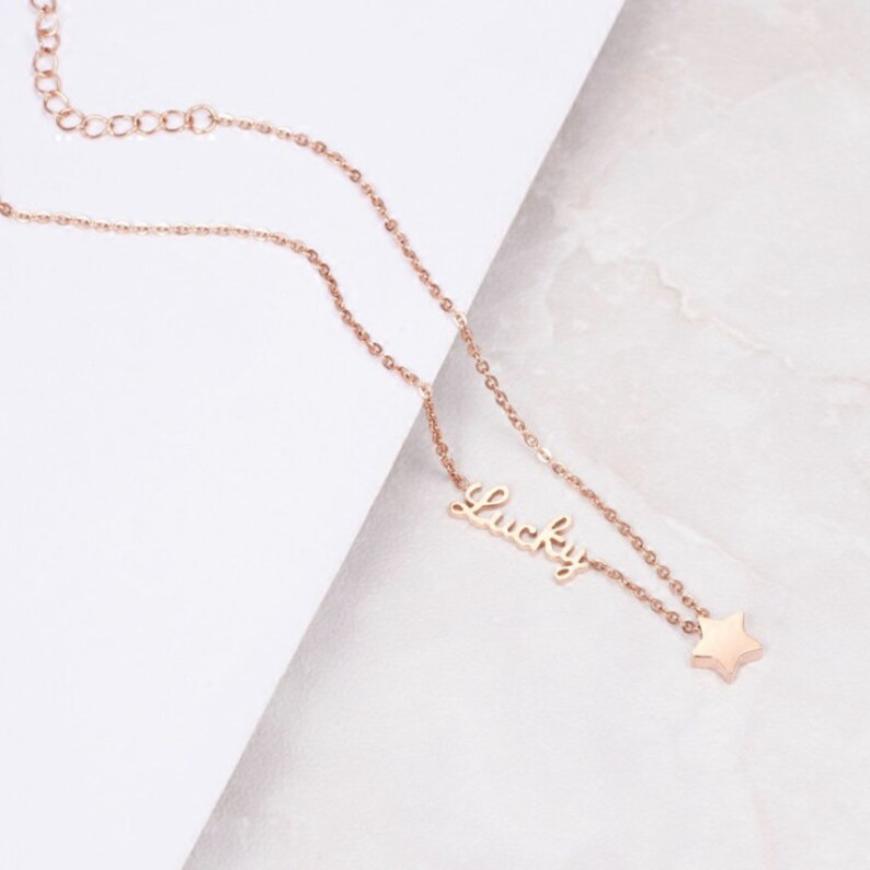 Puede incluir: Un collar de oro rosa con una cadena delicada y un colgante que dice "Lucky" con un peque&ntilde;o dije de estrella.