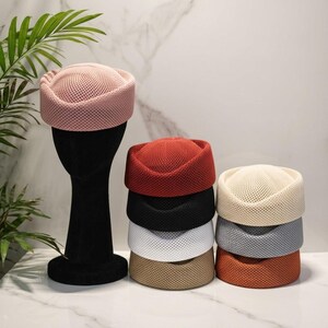 May include: A collection of brimless hats featuring a textured mesh construction. One light pink hat is displayed on a black mannequin head, while a stack of seven additional hats in red, black, white, beige, cream, gray, and orange are arranged nearby.