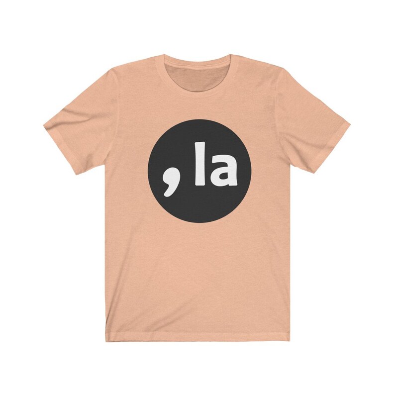 May include: A peach-colored t-shirt with a black circle containing a white comma and the word "la" in white.
