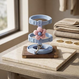 May include: A light blue pet collar and leash set. The set includes two collars, a leash, and a decorative pink and blue flower. The set is displayed on a wooden base with a marble top.