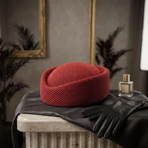May include: A round, rust-colored pillbox hat with a textured, woven knit pattern. The hat is displayed alongside a pair of smooth black leather gloves and a clear glass perfume bottle with a gold cap, all resting on a dark fabric drape.