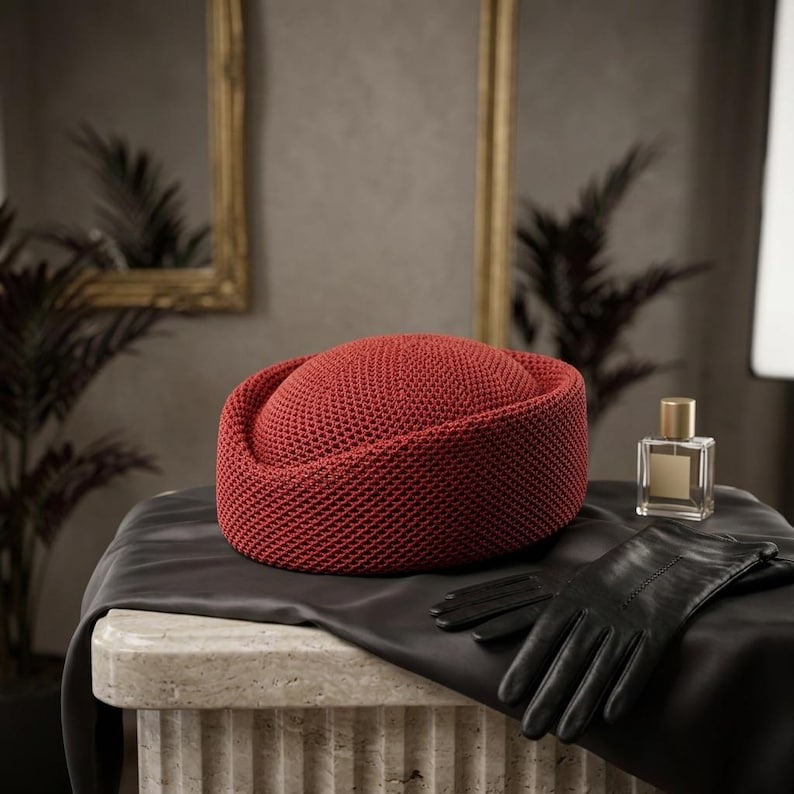 May include: A round, rust-colored pillbox hat with a textured, woven knit pattern. The hat is displayed alongside a pair of smooth black leather gloves and a clear glass perfume bottle with a gold cap, all resting on a dark fabric drape.
