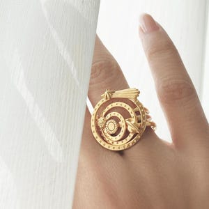 May include: A gold-colored ring with a circular design featuring concentric circles and intricate details. The ring is worn on a finger, with a white curtain in the background. The ring's design includes a small, rectangular element.