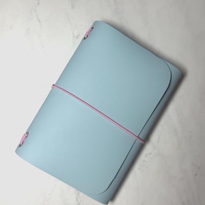 May include: A light blue leather journal with a pink elastic band closure. The journal has rounded corners and is closed. The edges of the journal are stitched with pink thread.