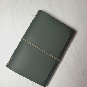 Könnte beinhalten: Ein dunkelgrünes Lederjournal mit abgerundetem Klappenverschluss. Ein beiges Gummiband umschließt das Journal, um es geschlossen zu halten. Die Ecken haben goldfarbene Metallakzente.