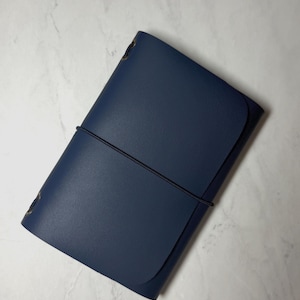 May include: A navy blue leather journal with a black elastic closure. The rectangular journal has rounded corners and brass-coloured eyelets. The cover is smooth and the journal is closed with a black elastic band.