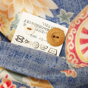May include: A white tag with Japanese text and care instructions, including a symbol for dry cleaning, a symbol for machine washing, and a symbol for tumble drying. The tag also includes the text "レーヨン 100%" and "ドライ セキュ系". There are two buttons, one white and one wooden, attached to the tag.