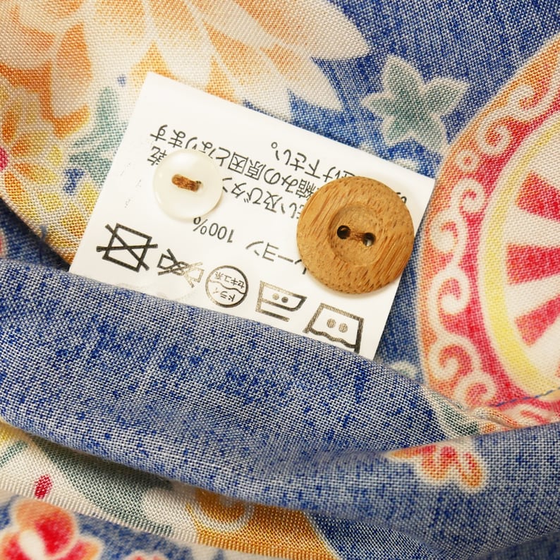 May include: A white tag with Japanese text and care instructions, including a symbol for dry cleaning, a symbol for machine washing, and a symbol for tumble drying. The tag also includes the text "レーヨン 100%" and "ドライ セキュ系". There are two buttons, one white and one wooden, attached to the tag.