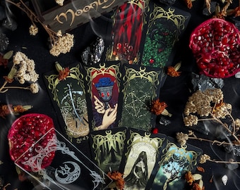 Op de afbeelding: Een spreiding van tarotkaarten met gouden accenten en ingewikkelde ontwerpen op een zwarte achtergrond. De kaarten zijn omringd door gedroogde bloemen, rook en granaatappels. De kaarten omvatten "De Aas van Zwaarden", "De Keizerin", "De Gehangene", "De Ster", "De Wereld" en "De Toren".